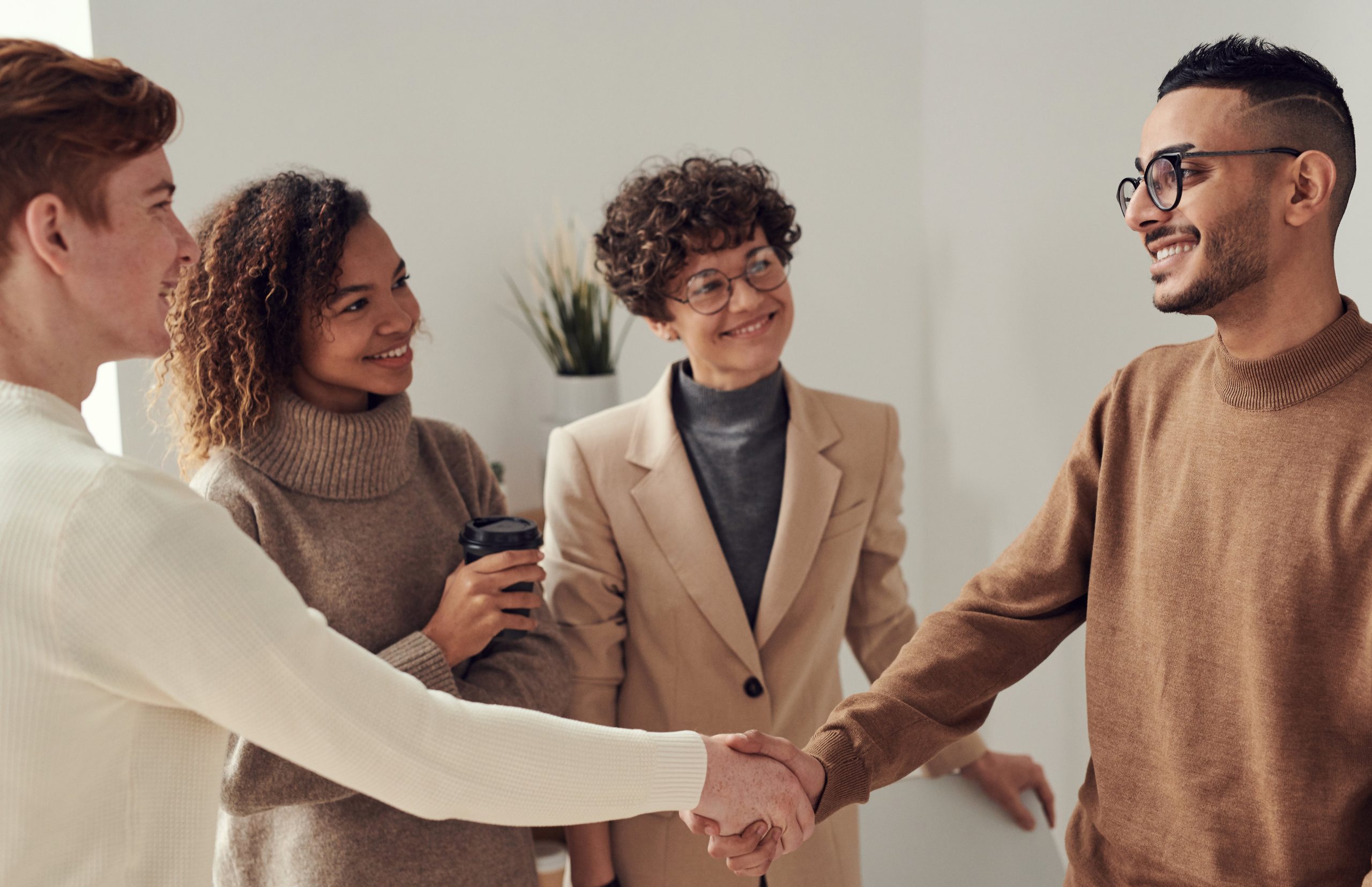 Four people smiling and shaking hands indoors, with one holding a coffee cup.