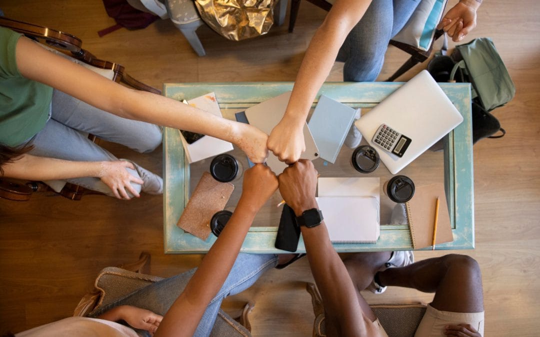 Four people sit around a table, doing a group fist bump over laptops, notebooks, and coffee cups.