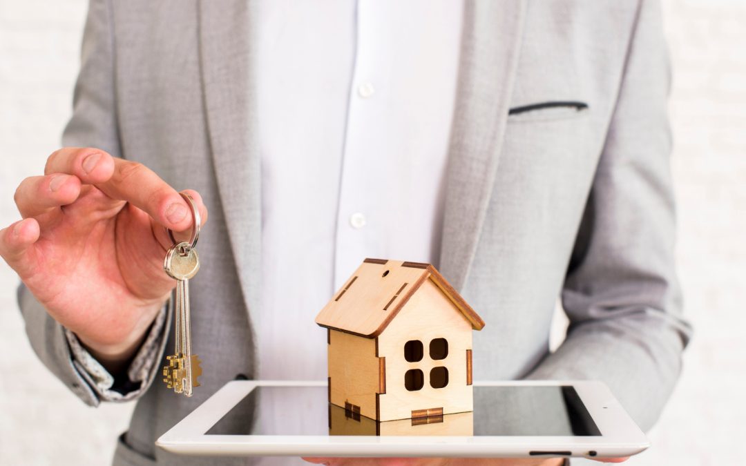 Person holding keys and a tablet with a small wooden house model, symbolizing real estate or renting.