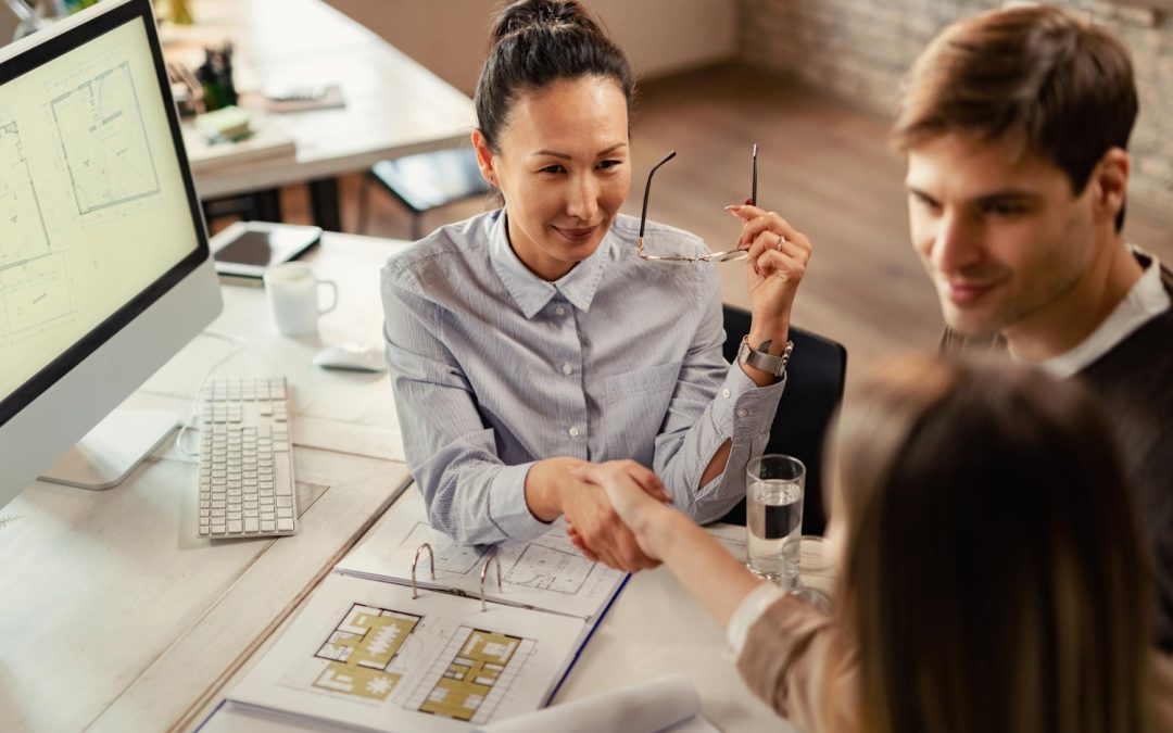 A woman smiles and shakes hands with a client across a desk with architectural plans on it.