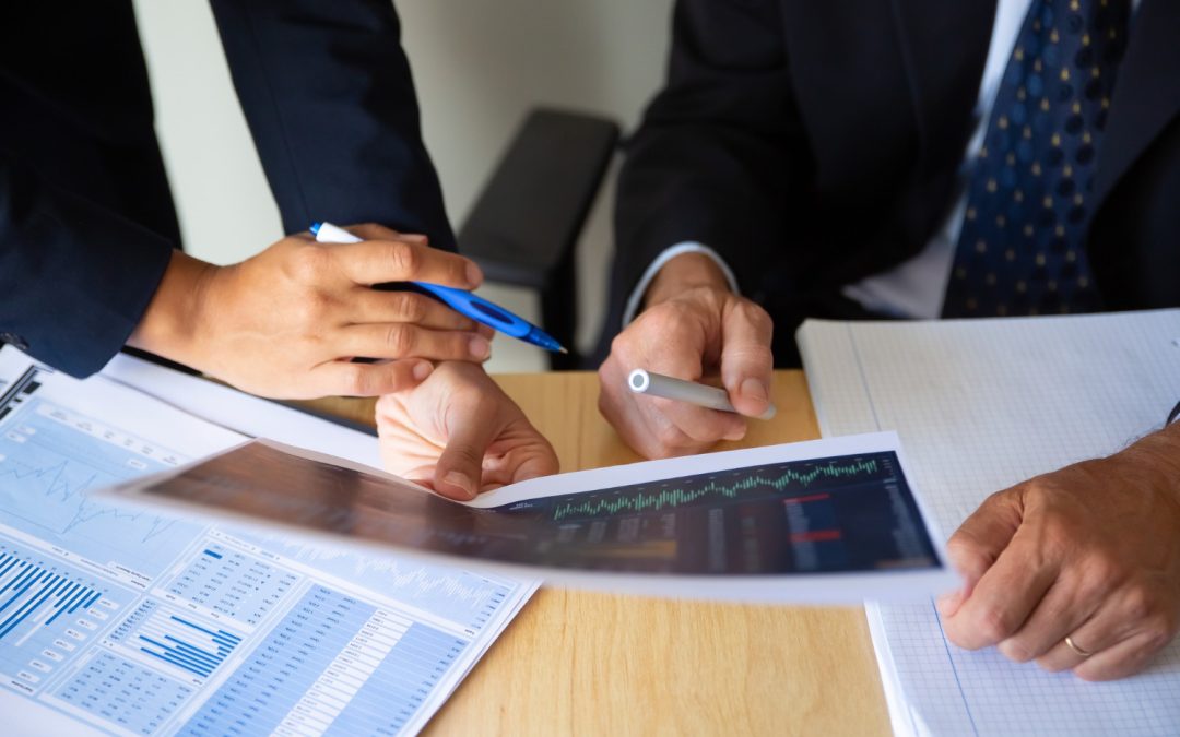 Two people in business attire discussing graphs and charts at a desk, pointing at documents with pens.