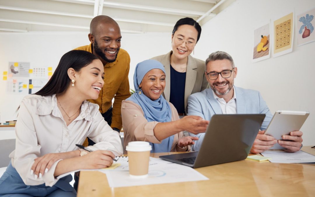 Five coworkers smiling and collaborating around a laptop and tablet in a bright office setting.