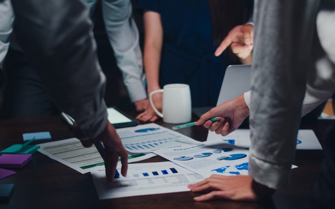 Hands of business people pointing at charts and graphs on a table during a meeting, with documents and a mug.